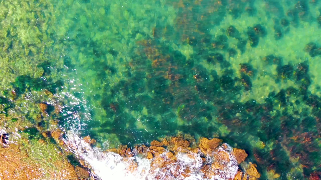 Top-down La Bocana shoreline view with a wider band of white foam over orange-brown rocks and transparent green water showing darker algae below the surface.