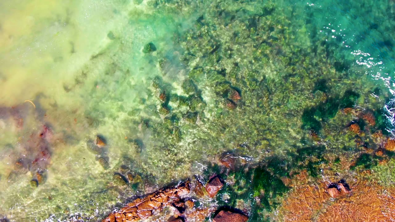Aerial view of clear green-blue water in La Bocana, Baja California Sur, with submerged rocks, darker algae patches, bright surface ripples, and no visible people.