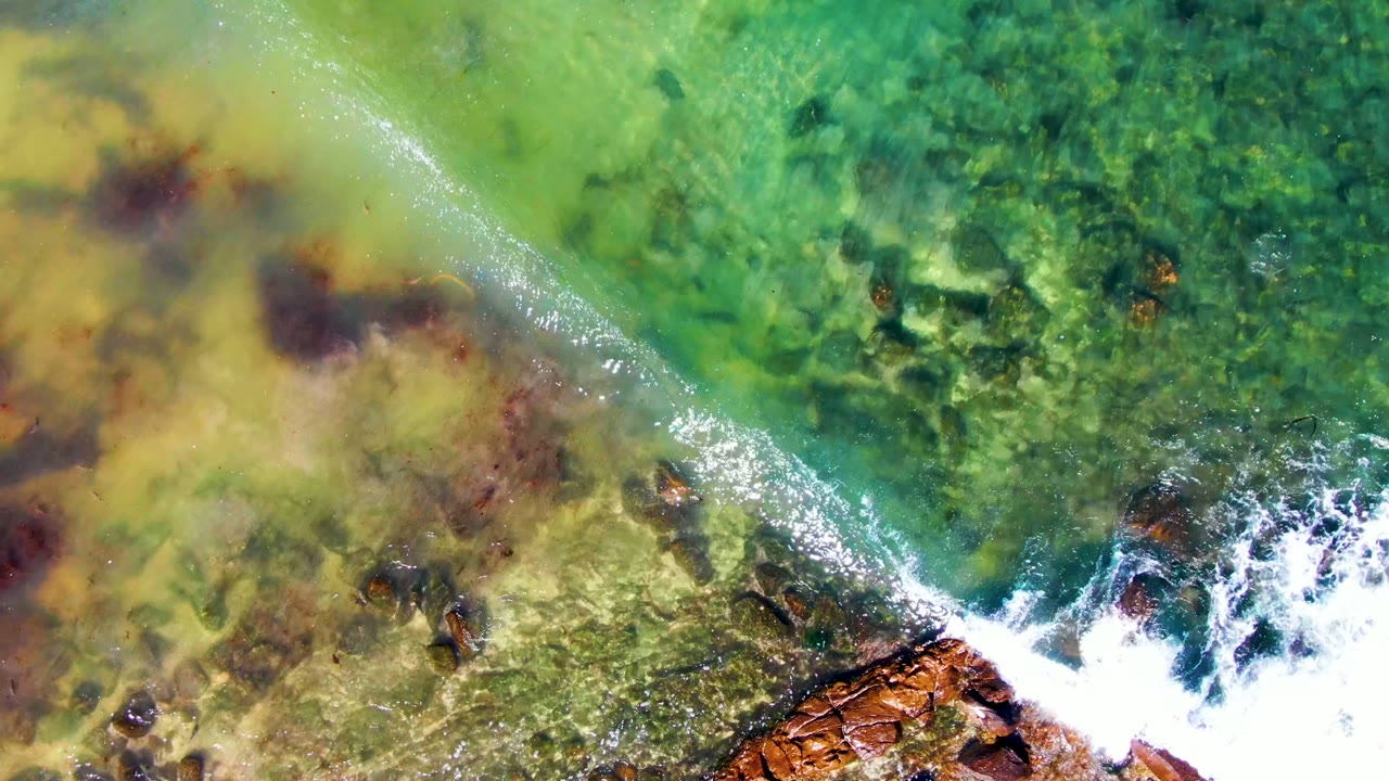 Overhead view of green water, submerged rocks, and a white wave breaking near the rocky shore at La Bocana, Mexico, with no visible people.
