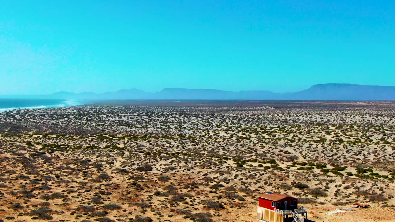 Early frame of La Bocana, Baja California Sur desert plain with scattered scrub, a small red-roofed structure near the lower center, distant mountains, blue sky, and no visible people.