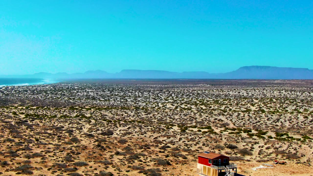 Wide desert plain with low vegetation, red-roofed structure, and hazy mountains.