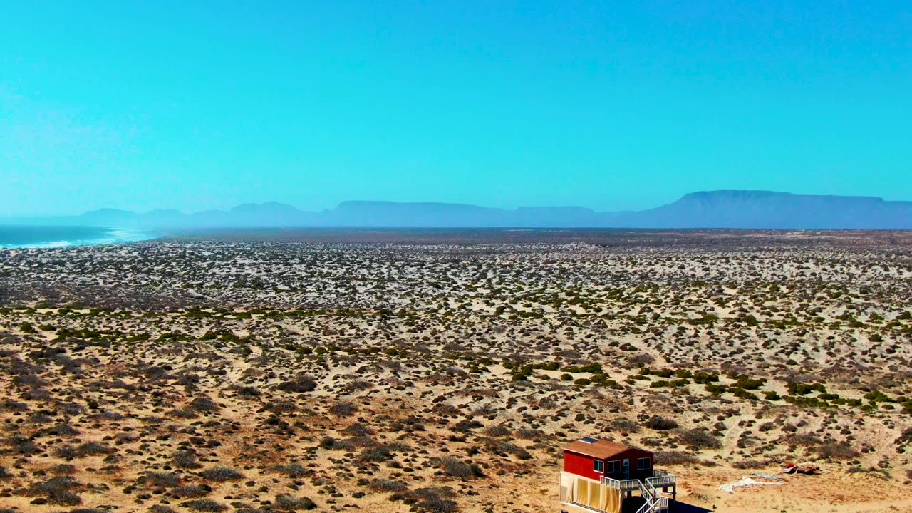 Middle early frame in La Bocana, Baja California Sur showing the small red-roofed building near the lower right, patchy scrub across the plain, mesa-like distant mountains, and no visible people.