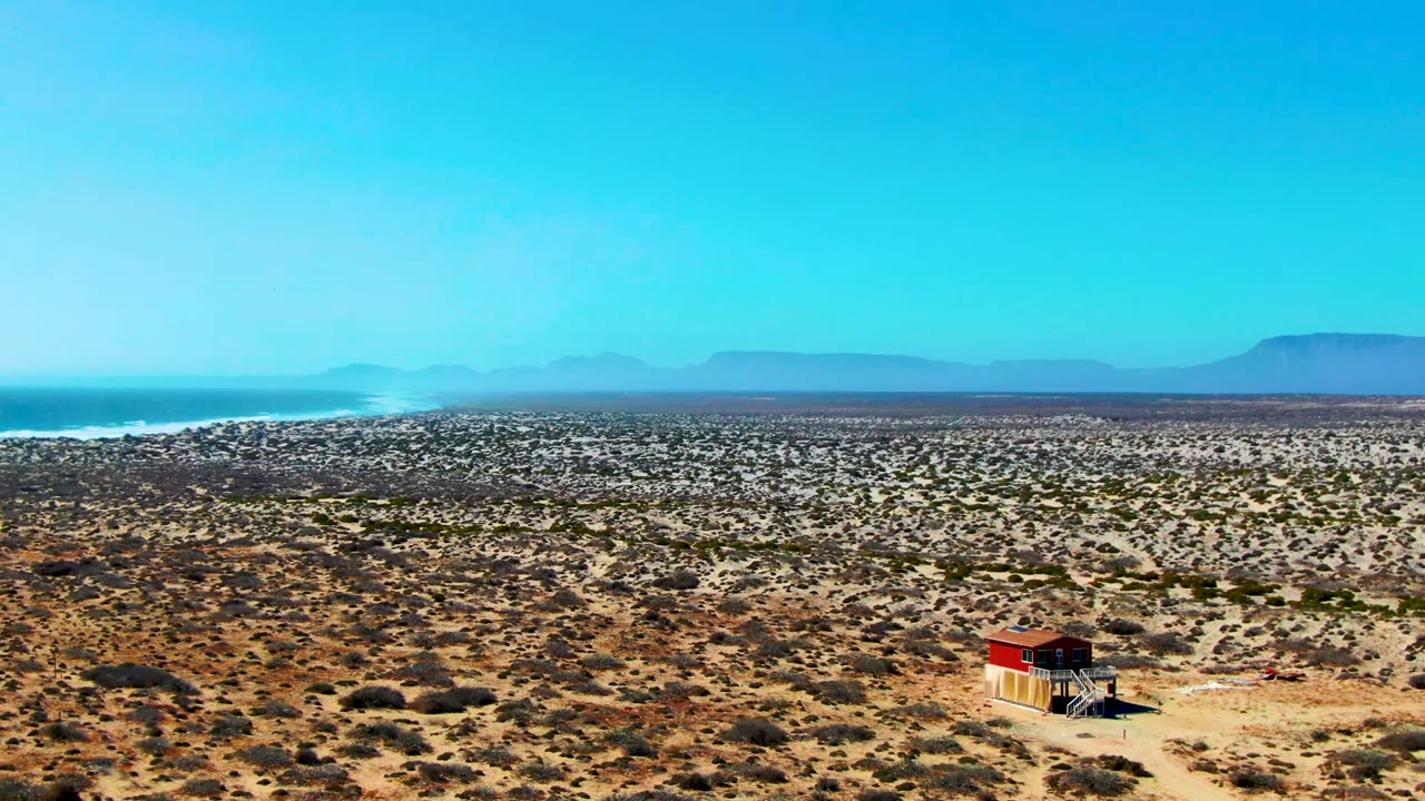At 0:00, a red-roofed hut sits low in the frame within the La Bocana desert, with the ocean on the left, distant mountains on the horizon, and no visible people.