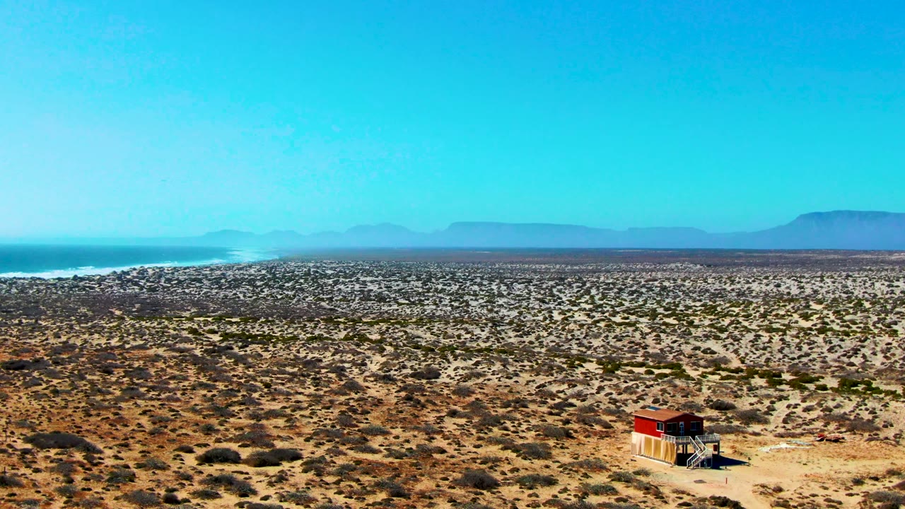 Aerial view of sparse desert scrub, a small red-roofed hut, ocean, and far mountains.