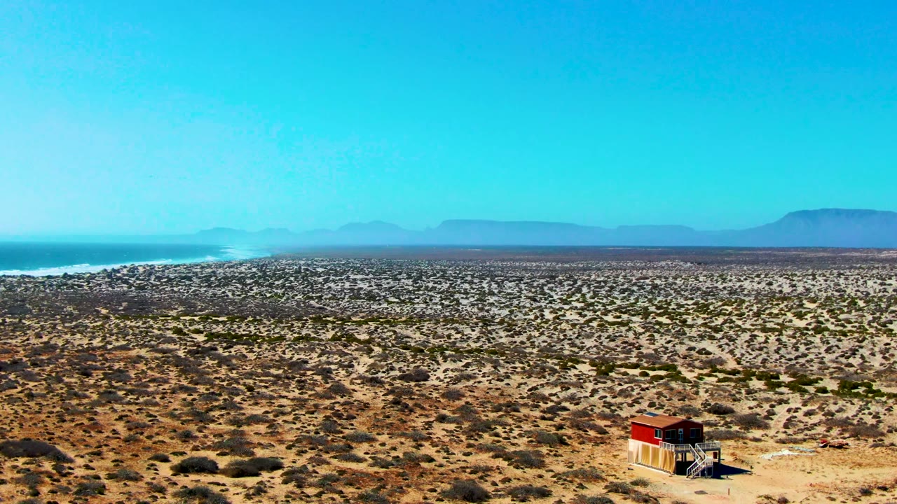 At 0:02, the hut has shifted toward the lower right of the La Bocana frame, with patterned desert scrub in front, ocean at left, mountains beyond, and no visible people.