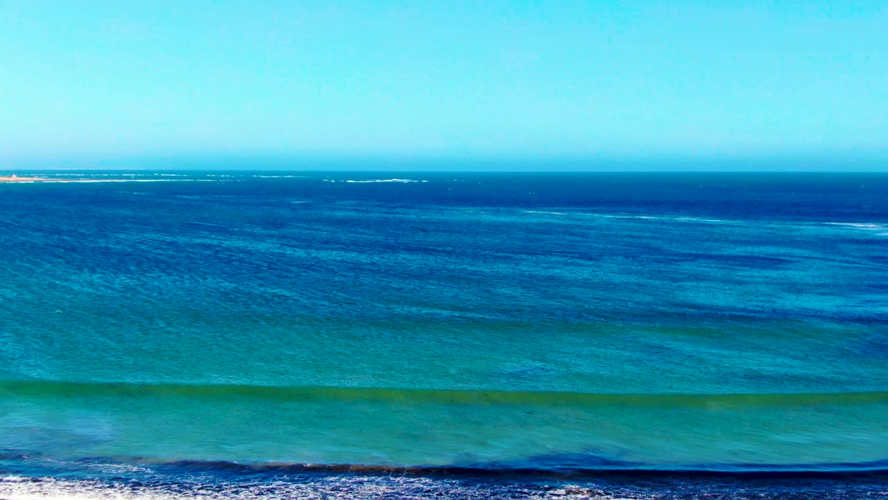 Aerial view of blue and turquoise water off La Bocana, Baja California Sur, with a low white surf line near the bottom edge and no visible people.