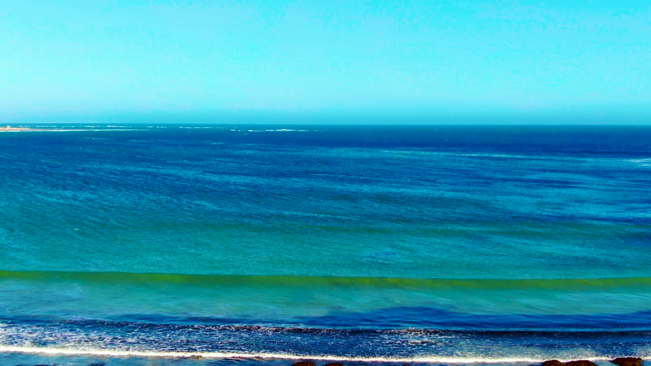 La Bocana shoreline from above, showing deep blue water beyond turquoise shallows, a white foam line at the bottom, and no visible people.