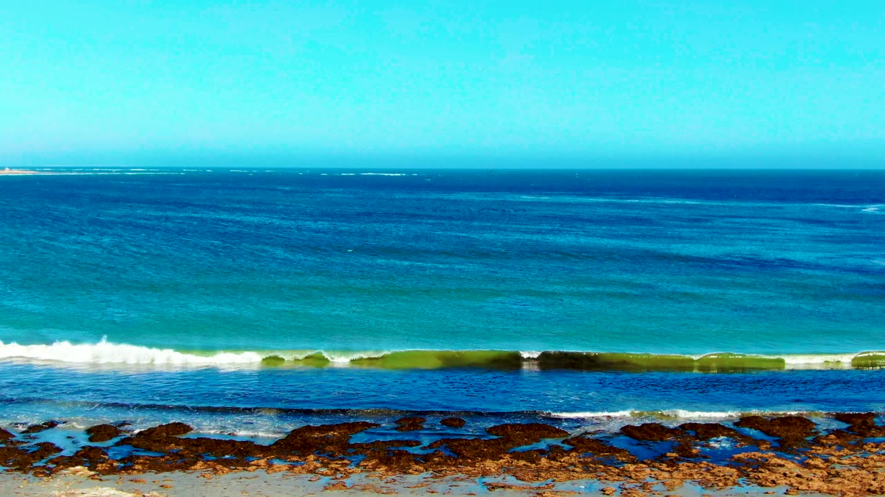 Wide elevated view of the La Bocana coastline with blue ocean bands, a white surf line, dark rocks along the shore, sandy foreground, and no visible people.