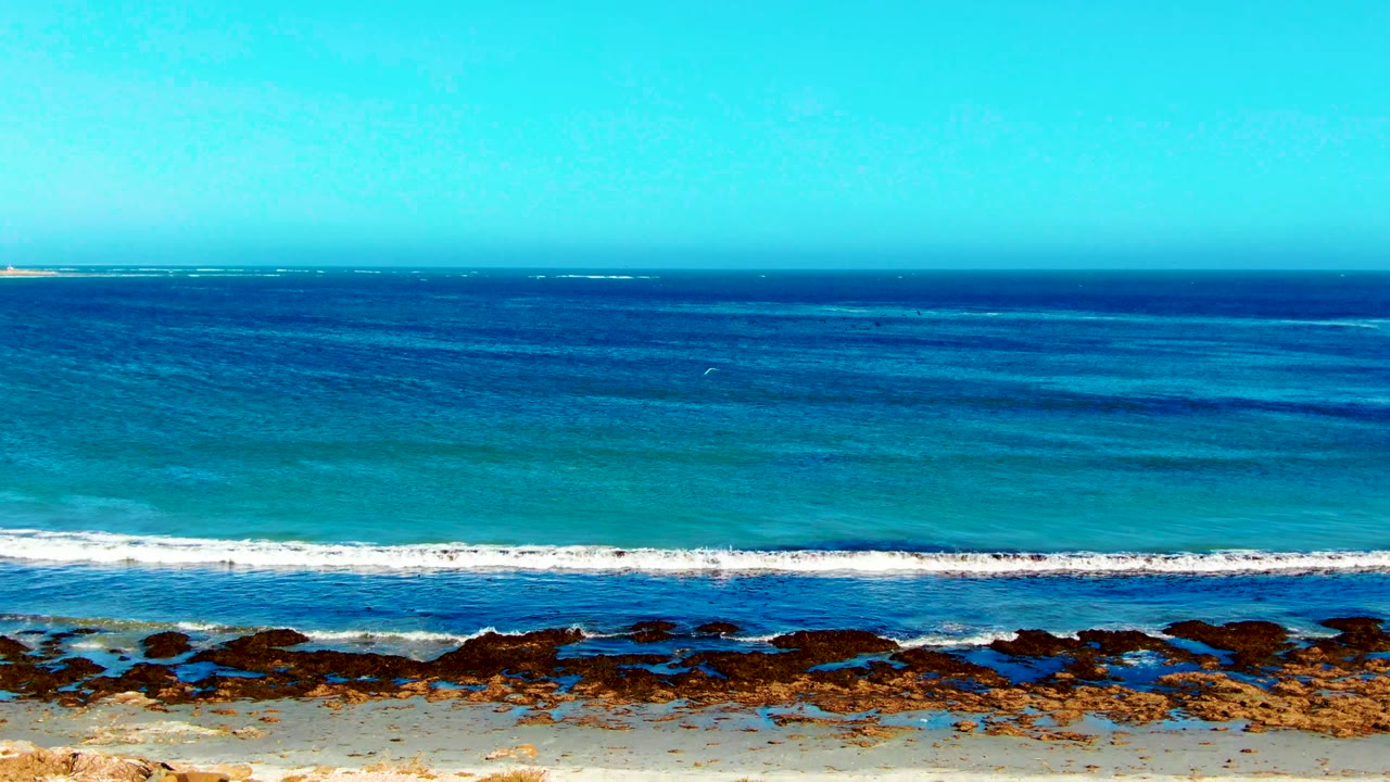 La Bocana shoreline frame with a long white wave breaking across the midground, deep blue water behind it, dark rocks at the waterline, pale sand below, and no visible people.