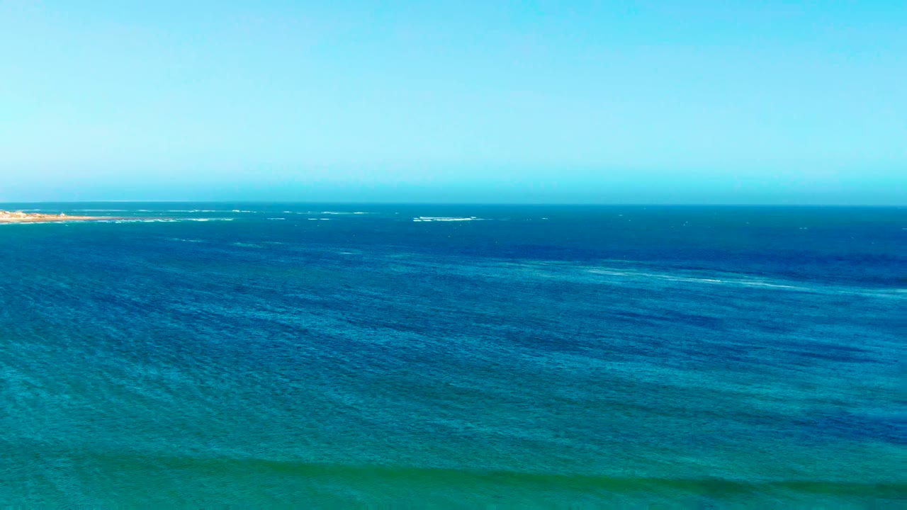 Wide aerial view of blue ocean off La Bocana, Baja California Sur, with a level horizon, turquoise shallows in the lower frame, thin whitecaps, and no visible people.