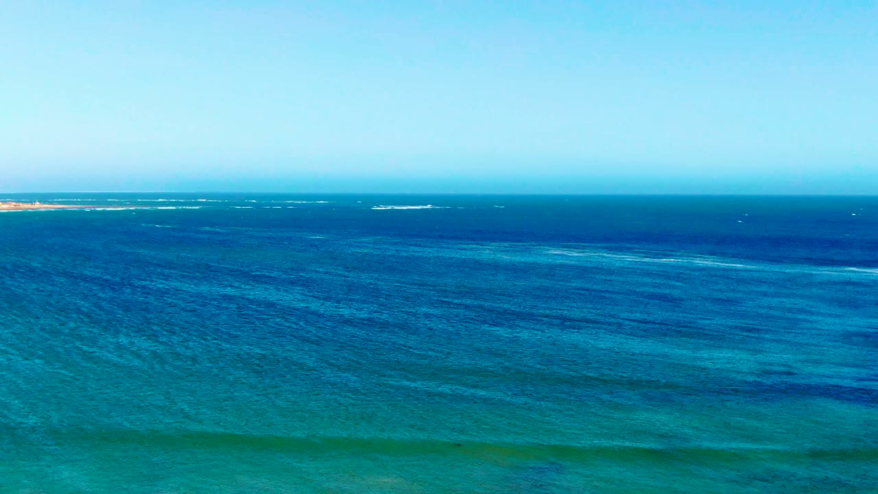 Wide drone-style view over La Bocana coastal water, where thin whitecaps sit near the horizon above textured blue water and turquoise shallows, with no visible people.