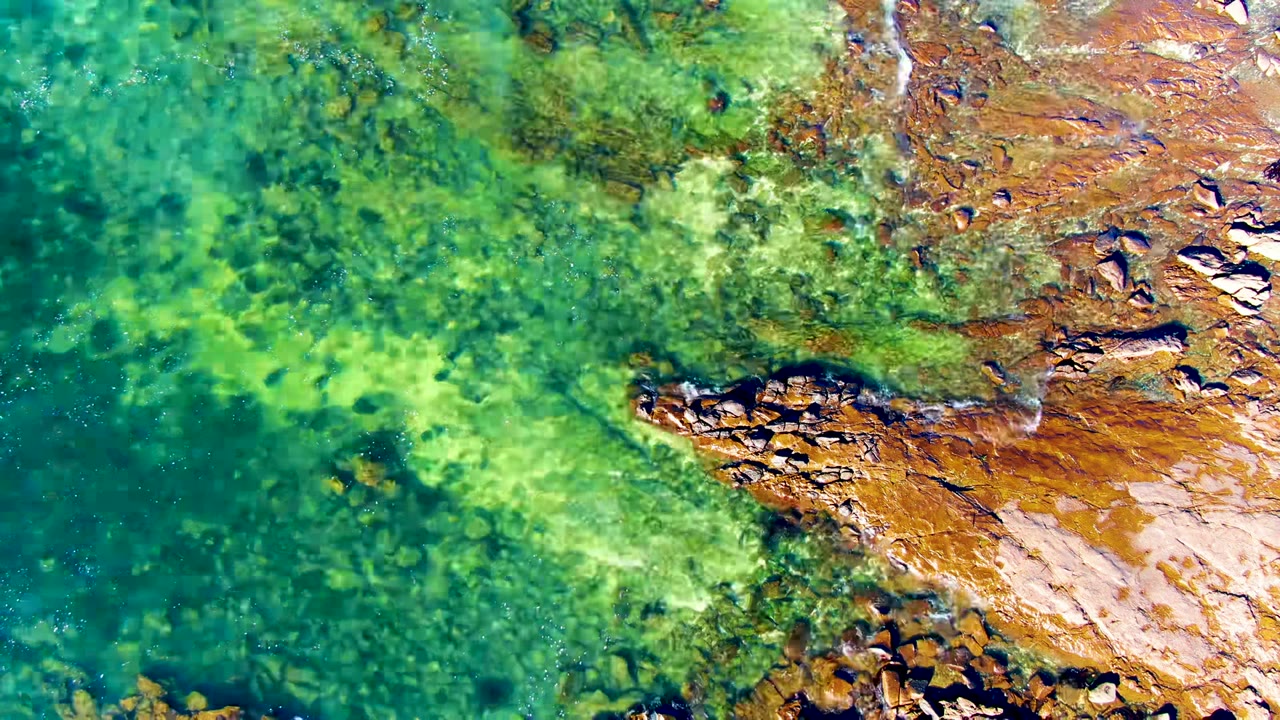 Overhead view of clear turquoise water and submerged green algae beside rust-colored rocks in La Bocana, Baja California Sur, with no visible people.