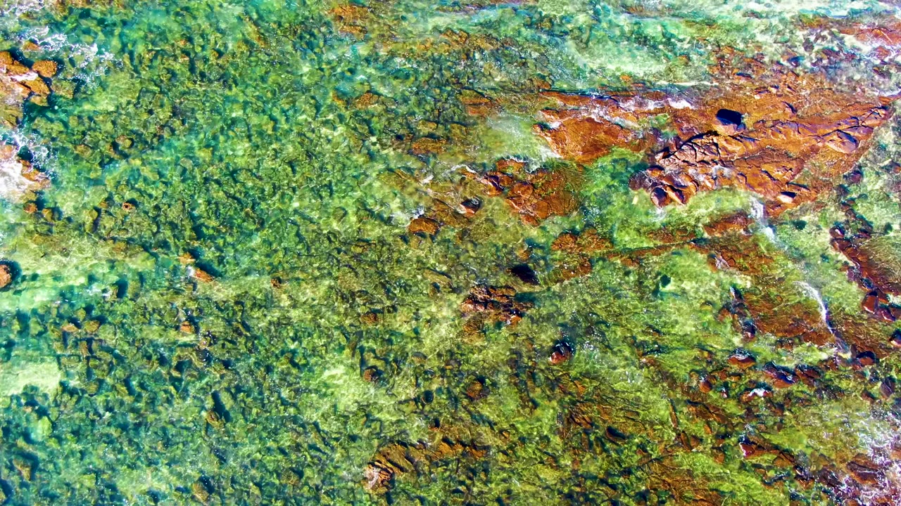 Top-down aerial view of clear green water and brown rocks at La Bocana, Baja California Sur, with white surf entering from the left and no visible people.
