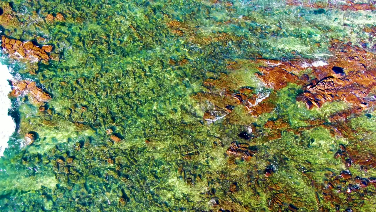 Top-down La Bocana coastal frame with mottled green water, rust-brown rock shelves, and a bright white foam streak cutting down the right side, with no visible people.