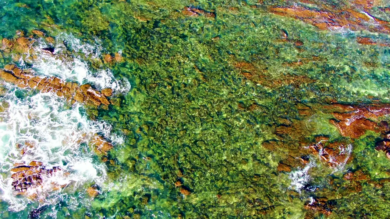 Overhead view of clear green-blue water washing across a rocky seabed in La Bocana, Baja California Sur, with white foam entering from the left and no visible people.