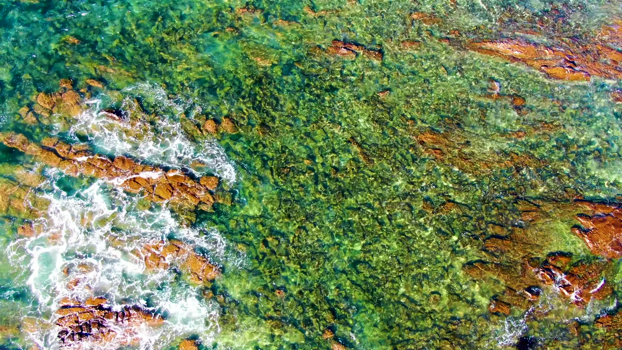 Overhead La Bocana frame with clear green water, scattered submerged rocks, and white foam clustered near the lower right edge, with no visible people.