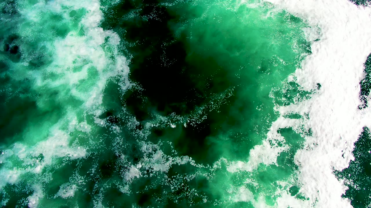 Overhead view of green ocean water at La Bocana, Baja California Sur, with white foam entering from the right and dark underwater patches visible, no visible people.