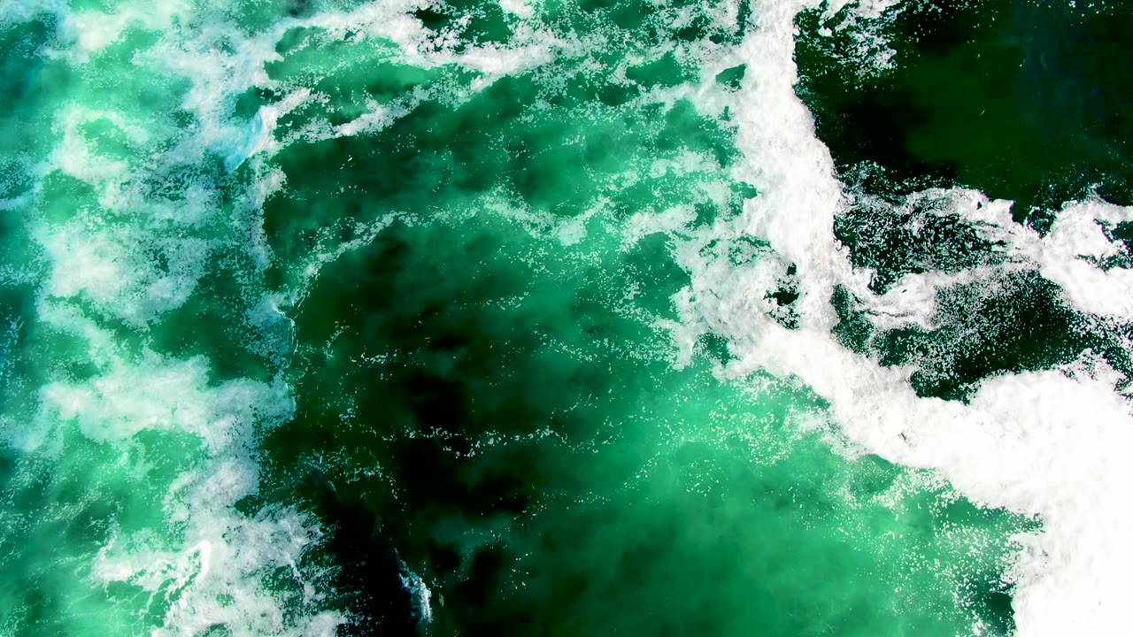 At 0:00, an overhead view in La Bocana, Baja California Sur shows green ocean water crossed by bright white surf foam, with dark underwater patches and no visible people.