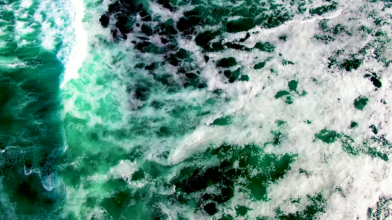Top-down view of turquoise and dark green surf with white foam spreading across the water in La Bocana, Baja California Sur, Mexico, with no visible people.