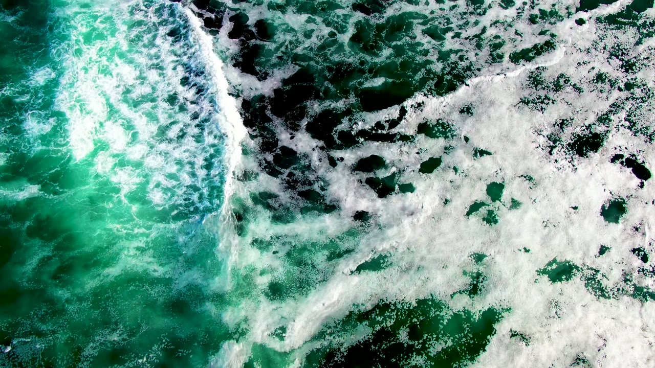 Overhead surf frame with a bright breaking wave line beside spreading white foam on green water in La Bocana, Baja California Sur, Mexico, with no visible people.