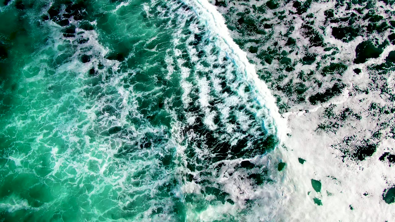 Overhead view of turquoise and green ocean water at La Bocana with white surf foam crossing the frame and no visible people.