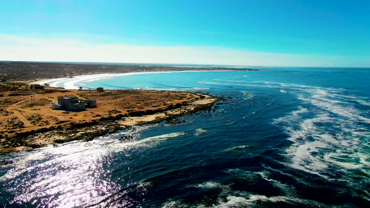 Opening aerial frame over La Bocana, Baja California Sur, with a low coastal building on dry land, dark rocky shore, bright surf, blue water, and no visible people.