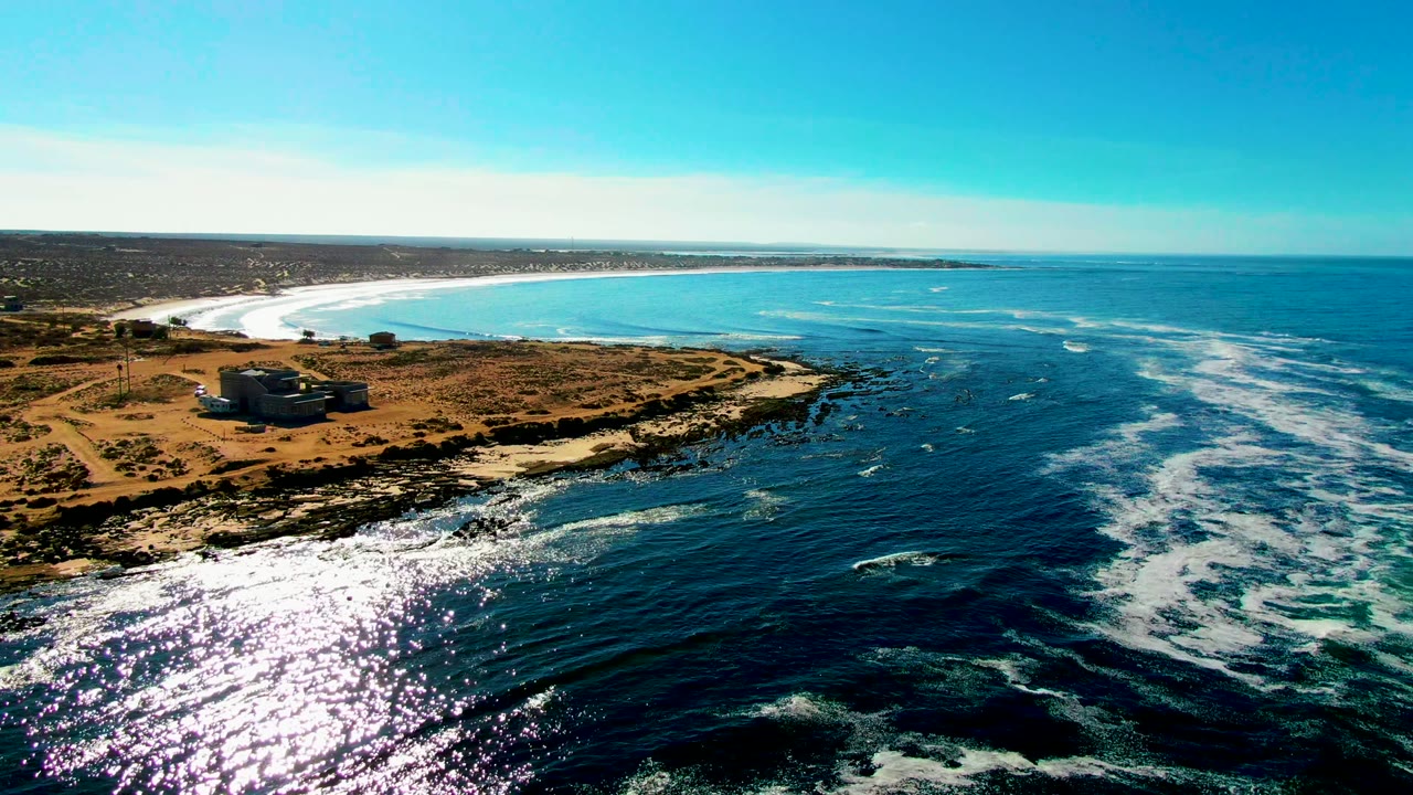 Aerial coastline with sun glare on waves near a shore building.