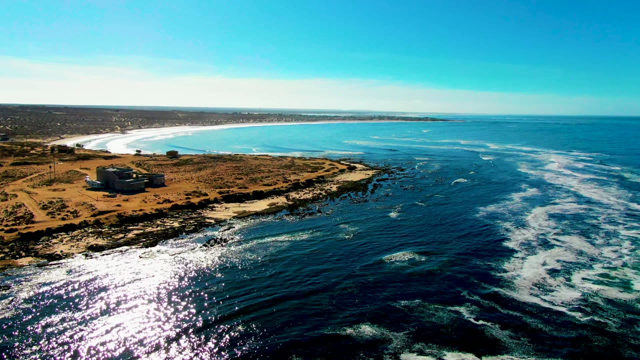 La Bocana aerial frame with the building on the left shore, a darker blue channel through the center water, white wave streaks to the right, and no visible people.