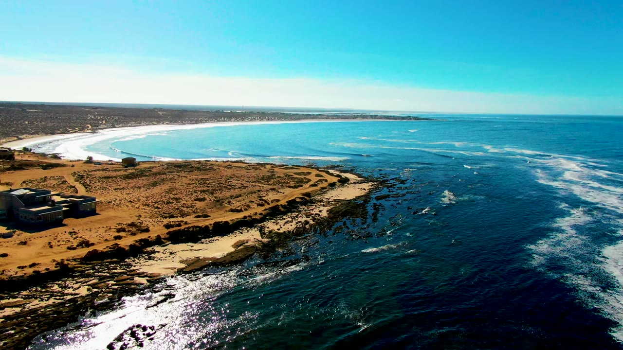 Opening aerial frame over La Bocana, Baja California Sur, with small structures at the left edge, tan coastal scrub in front, a curved sandy beach beyond, blue ocean to the right, and no visible people.