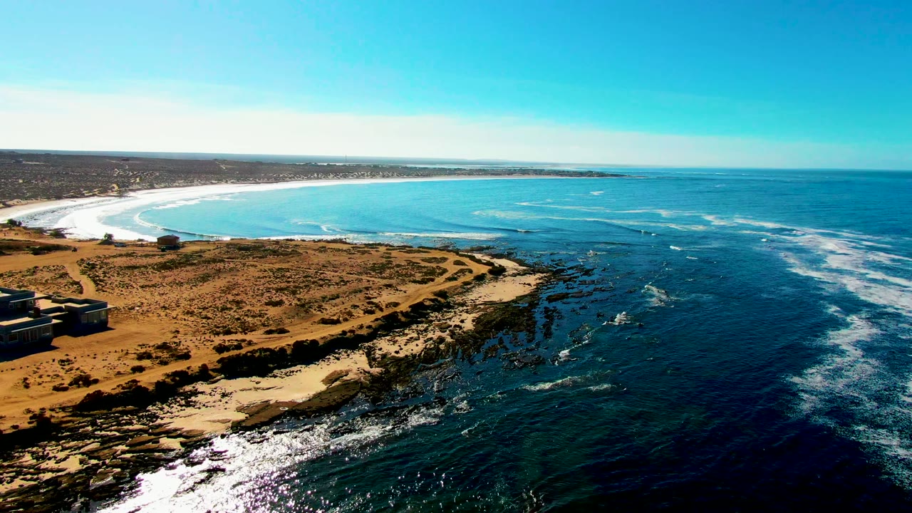 Drone view of sandy bay, rocky point, scrubland, and ocean surf.