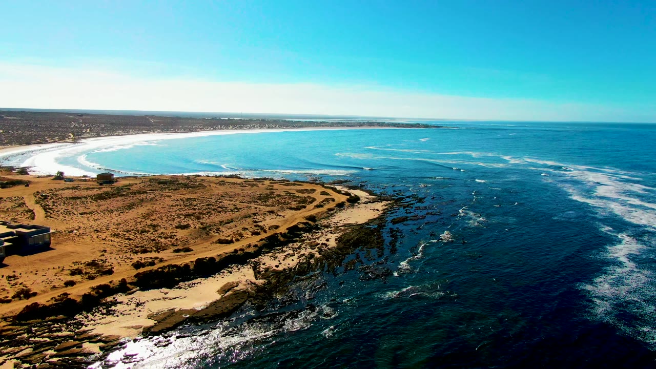 At 0:02, the La Bocana aerial holds a wide composition of tan land and dark rock ledges in the foreground, a pale sandy beach curving across the background, blue ocean to the right, and no visible people.
