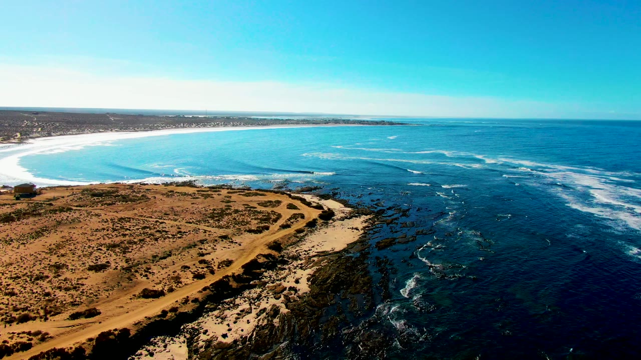 Aerial frame over La Bocana, Baja California Sur showing dry brown terrain, a sandy path, rocky shoreline, blue ocean, white surf, and no visible people.