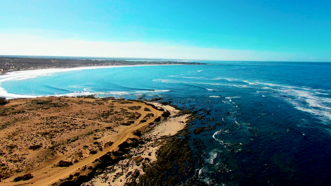 Wide aerial coastline with turquoise bay, rocky shore, and dry land in foreground.
