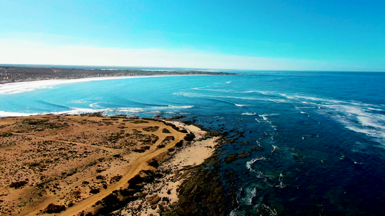 La Bocana aerial frame showing the rocky point below, dark reef-toned water near shore, white surf lines offshore, dry land to the left, and no visible people.