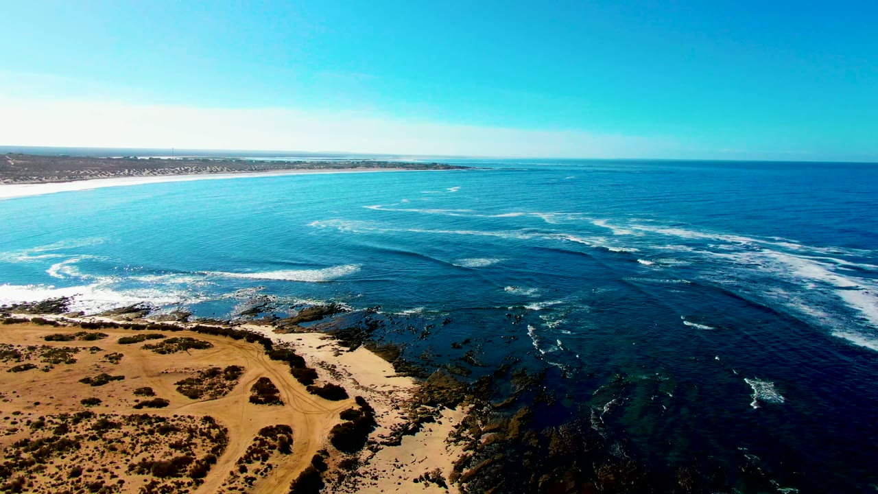 At La Bocana, Baja California Sur, an aerial view shows sandy dunes in the lower left, dark rocky reef below, blue ocean beyond, and no visible people.