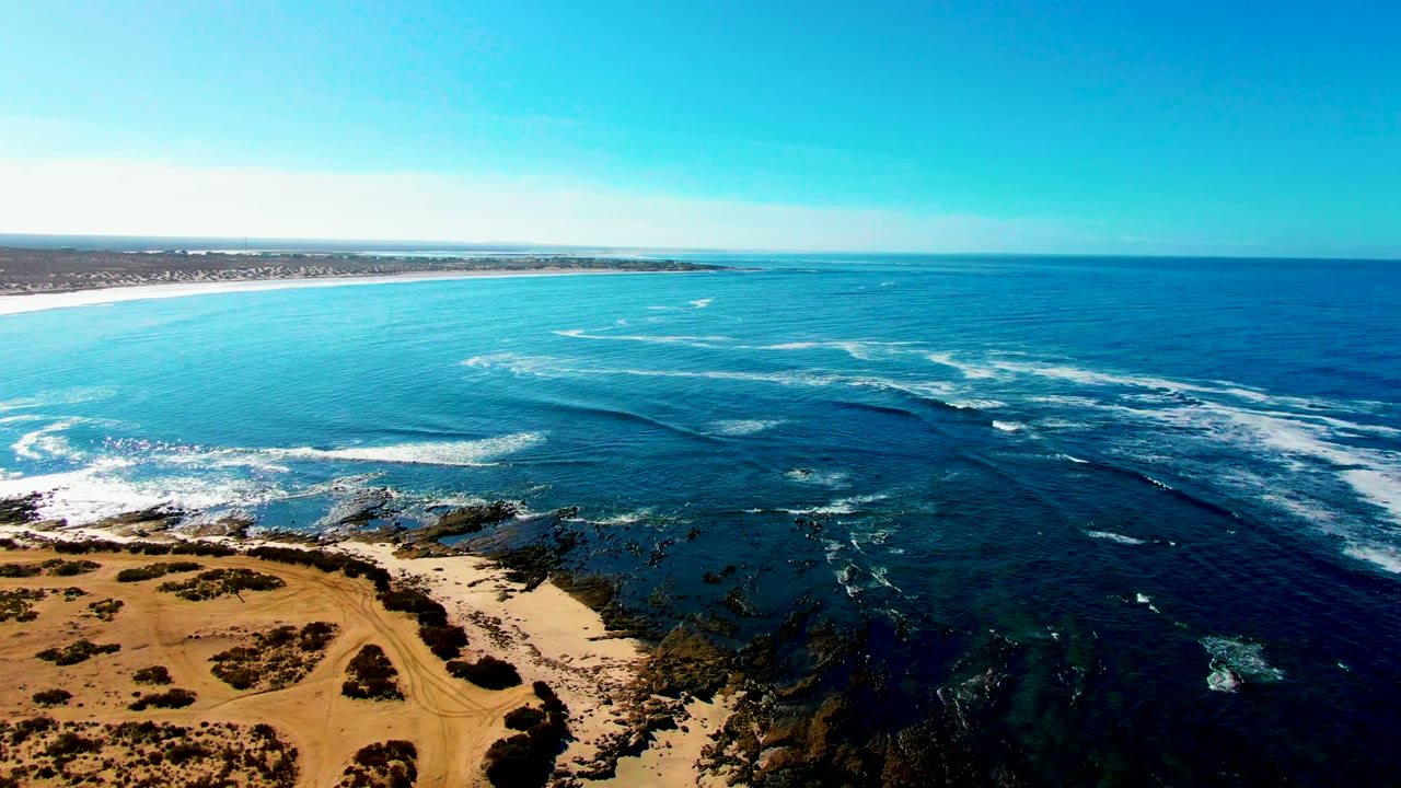 Wide drone view of turquoise water and white surf lines along a rocky shoreline.