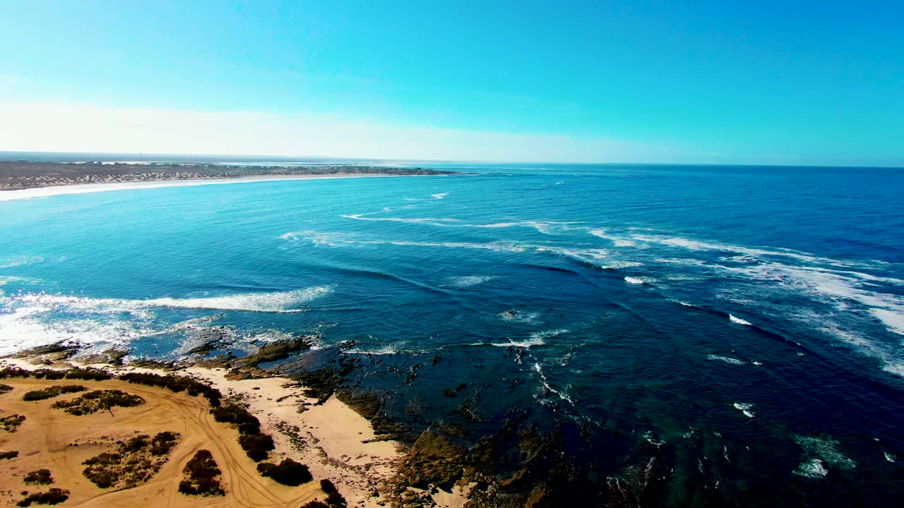 At La Bocana, Baja California Sur, the drone view holds the sandy dune edge at lower left while bands of foam cross the dark reef and open ocean, with no visible people.