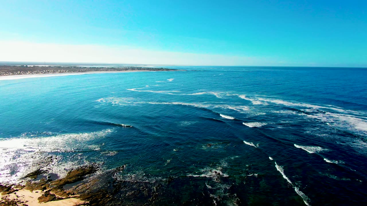 High aerial view of blue ocean off La Bocana, Baja California Sur, with white waves breaking over dark rocky shallows, sandy coast at left, clear sky above, and no visible people.