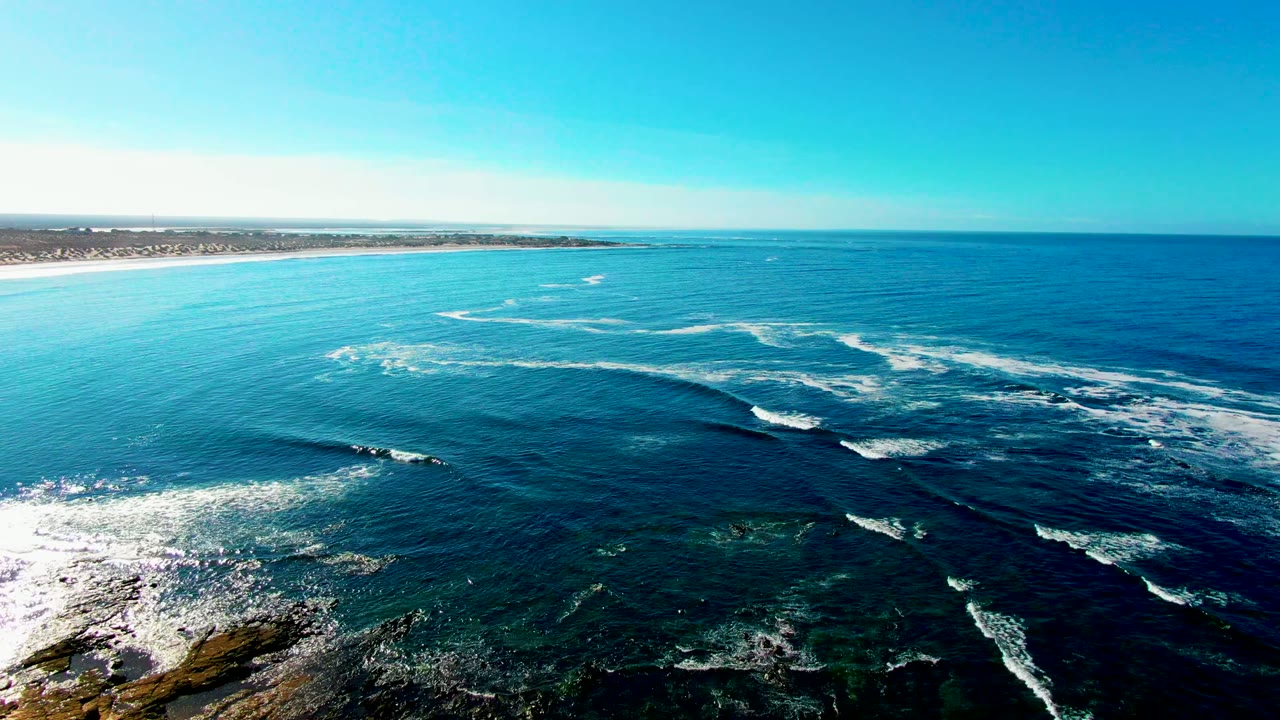 White foam spreads across rocky shallows beside a sandy coast