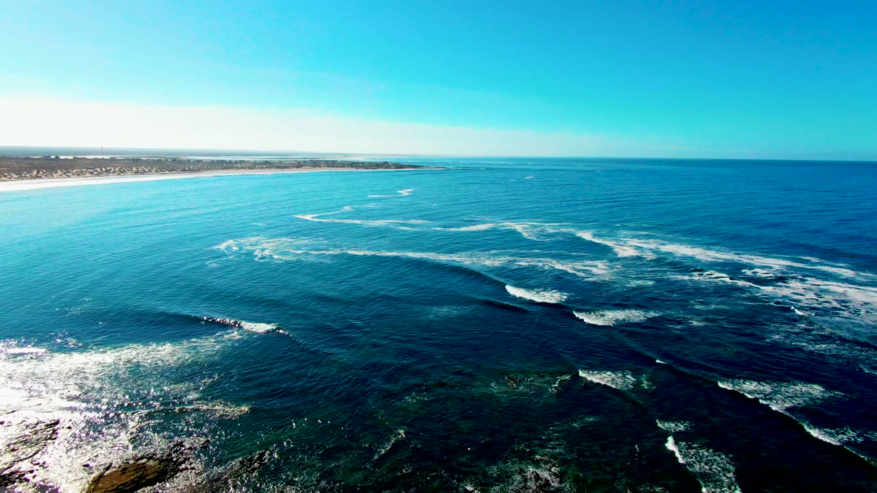 Wide drone view at La Bocana with long white surf bands curving through blue water, darker reef patches below, a low sandy coastline at left, clear horizon, and no visible people.