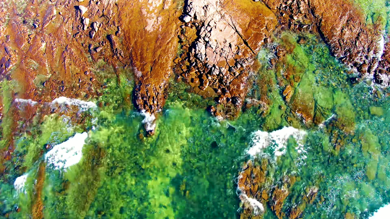 Top-down aerial frame of clear green water washing against brown rocks at La Bocana, Baja California Sur, with white foam along the shore and no visible people.