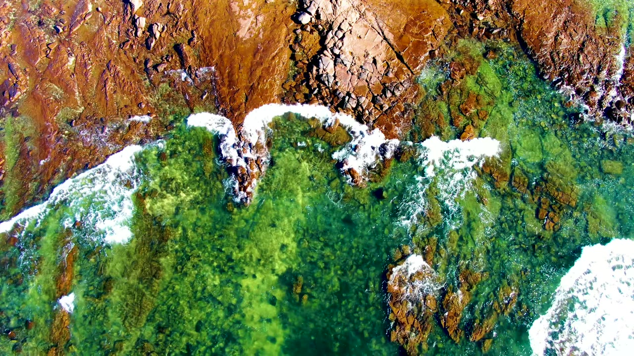 Overhead frame of La Bocana coastline with brown rock slabs along the top and transparent green-blue water below, no visible people in frame.