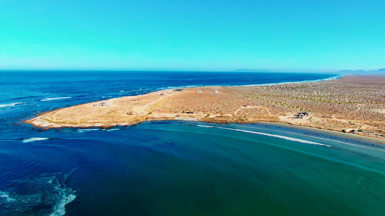 Opening aerial frame over La Bocana, Baja California Sur, with a sandy peninsula, sparse vegetation, white surf lines, deep blue water, turquoise shallows, and no visible people.