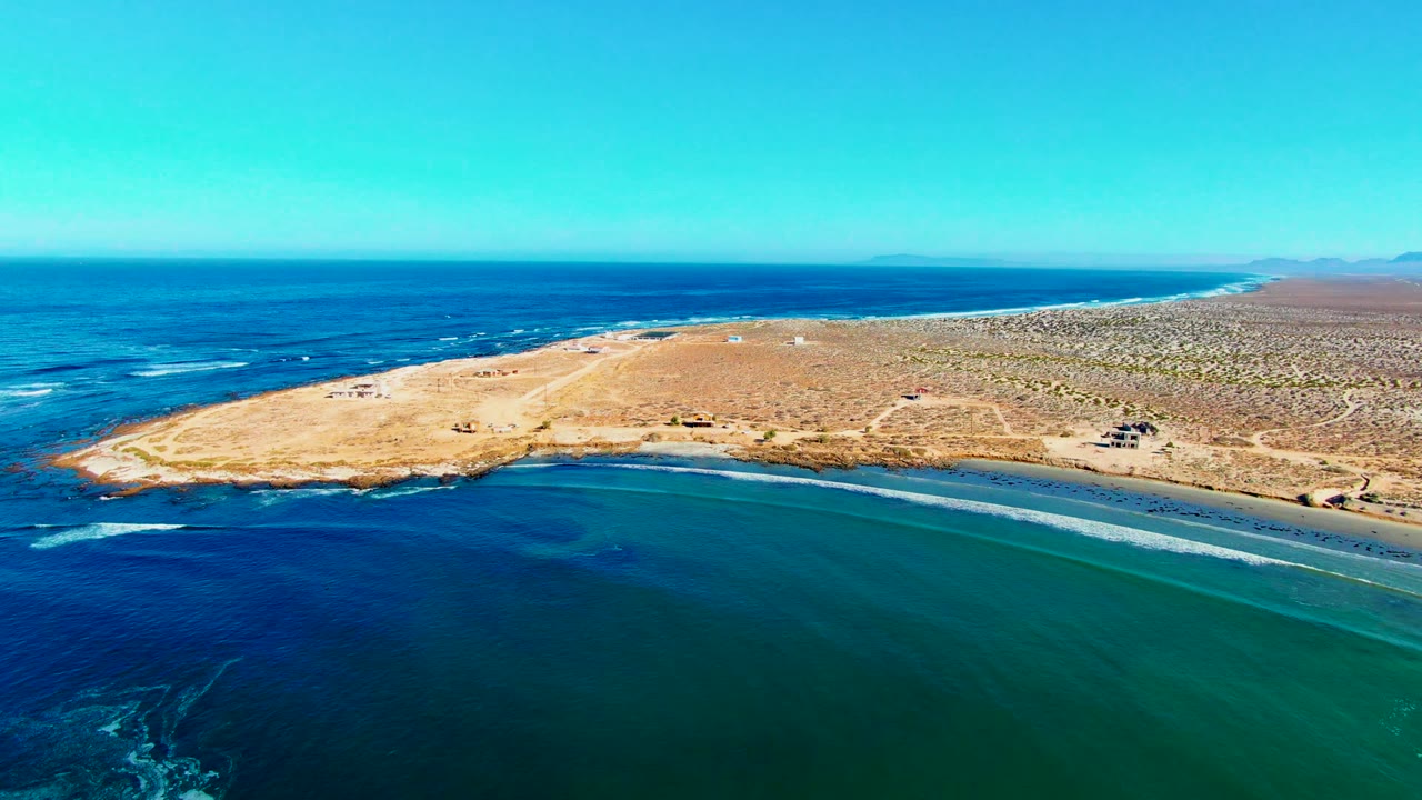 La Bocana aerial view with the peninsula crossing the frame, dark blue ocean on the left, green-blue shallows on the right, a distant coastline, and no visible people.
