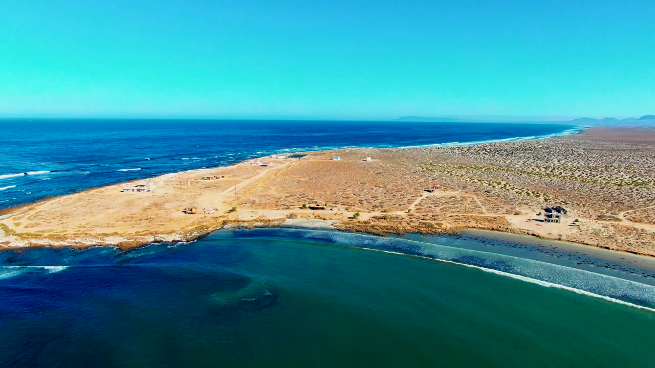 Aerial frame over La Bocana, Baja California Sur, showing a green-blue cove in front, sandy desert shoreline behind it, open ocean beyond, and no visible people.