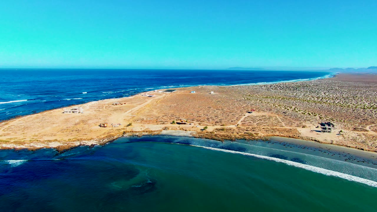 Wide aerial frame over La Bocana showing the cove water in the foreground, white wave bands near shore, dry tan land stretching inland, distant mountains on the horizon, and no visible people.