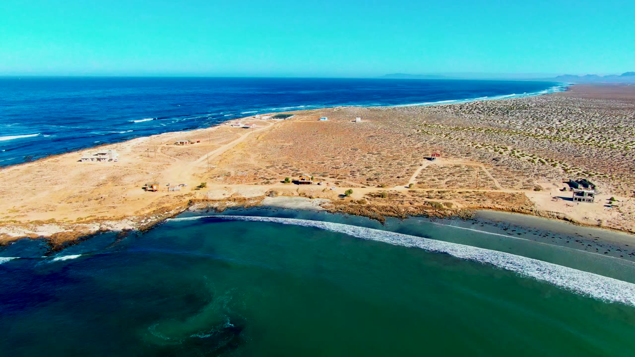 Aerial frame of La Bocana showing green inlet water below a sandy shore, dry scrubland, distant Pacific surf, and no visible people.