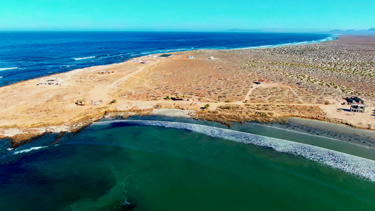 Aerial frame of La Bocana where a white surf line crosses the green inlet, with sandy shore, sparse desert vegetation, dirt tracks, and no visible people.