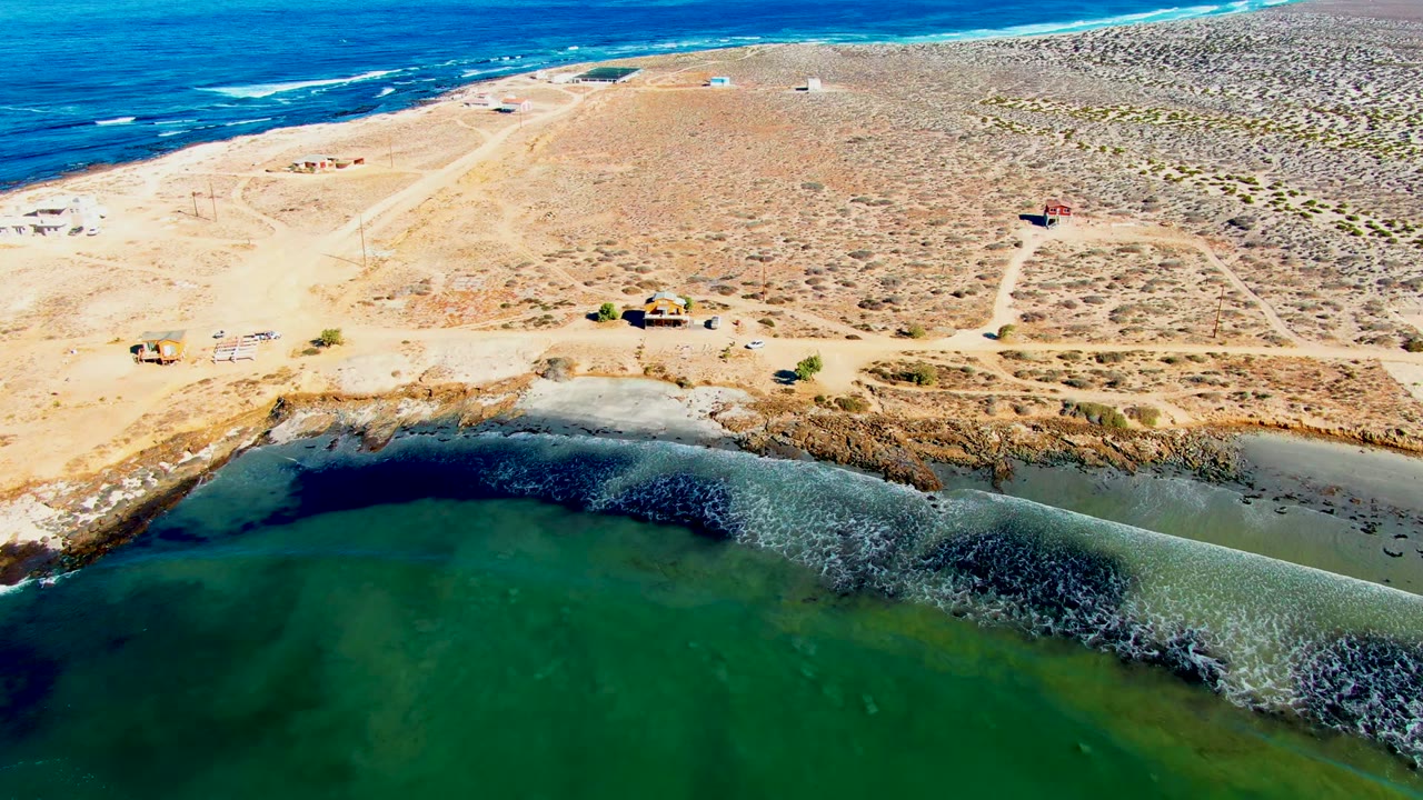 Aerial frame over La Bocana showing green lagoon water in the foreground, a rocky tan shoreline, sandy dirt roads, sparse structures, blue ocean at the top, and no visible people.