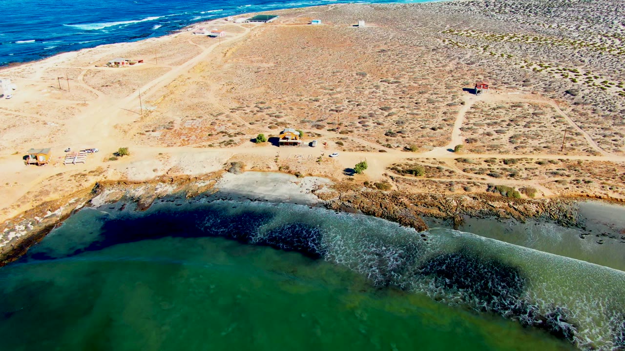 Drone view of green coastal water, tan shoreline, dirt tracks, vehicles, and small buildings.
